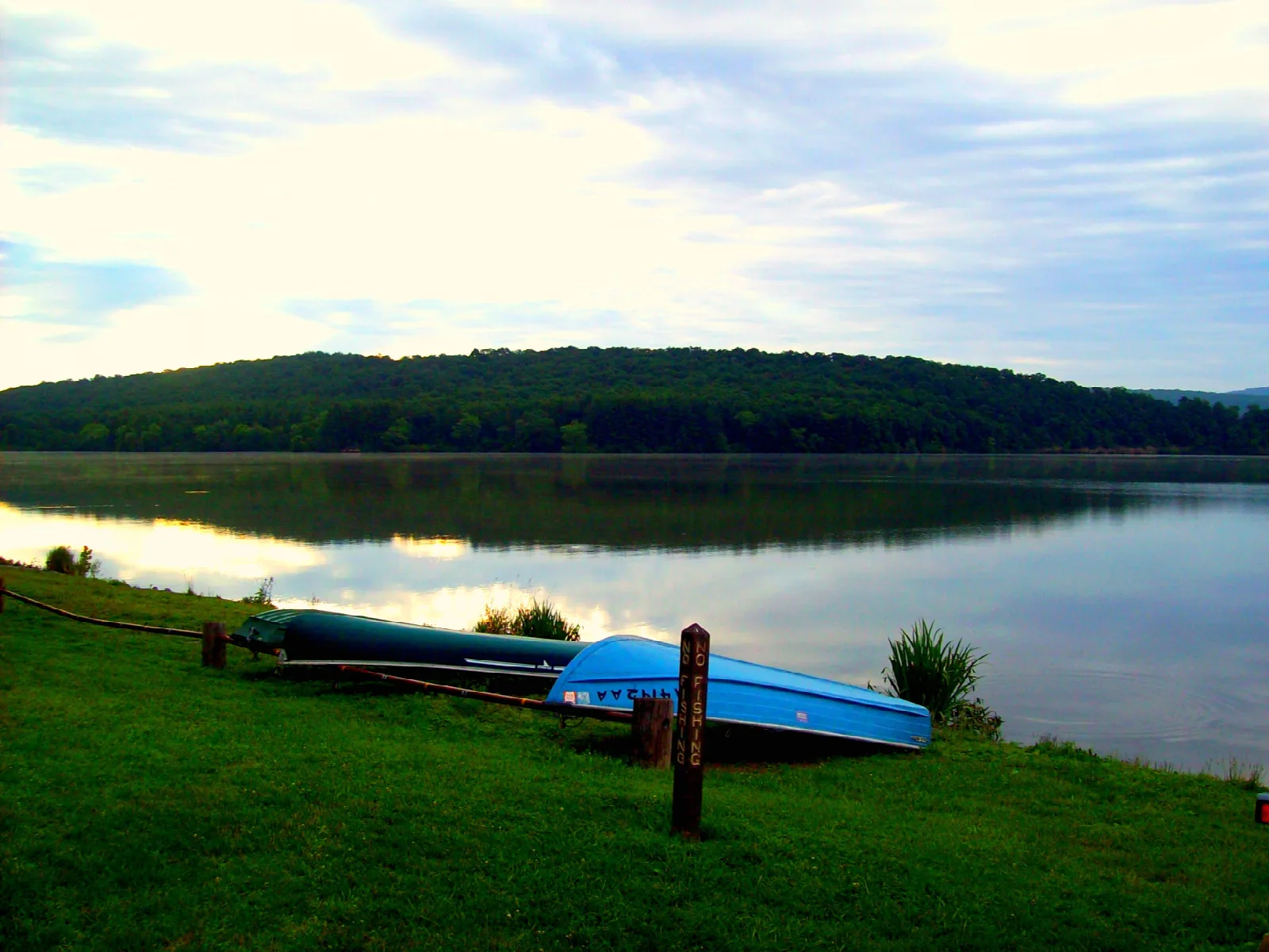 Calm lake reflecting a forested hill, with two small boats on the grassy bank