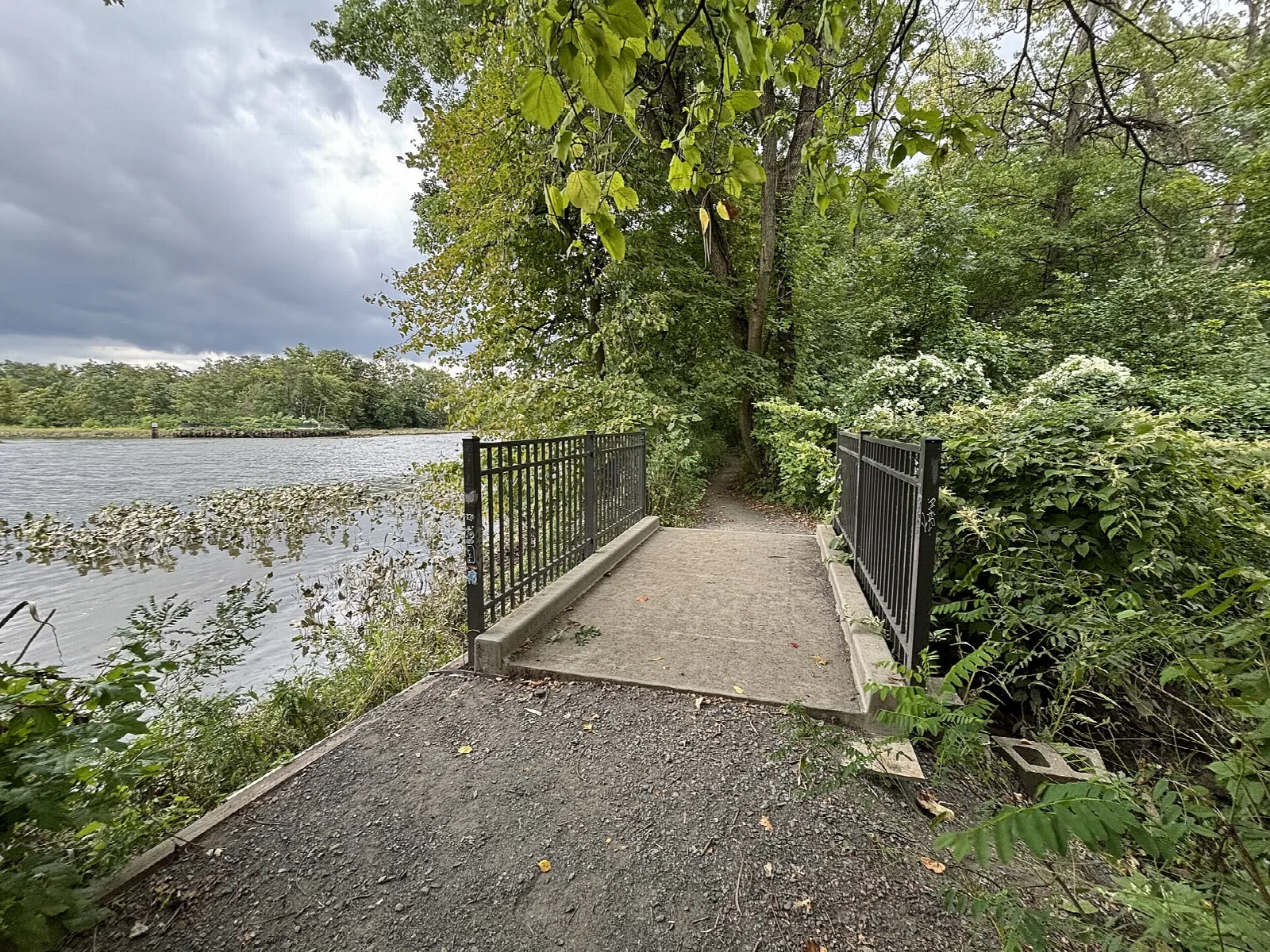 Gravel path and small bridge with metal rails beside the water, surrounded by green trees