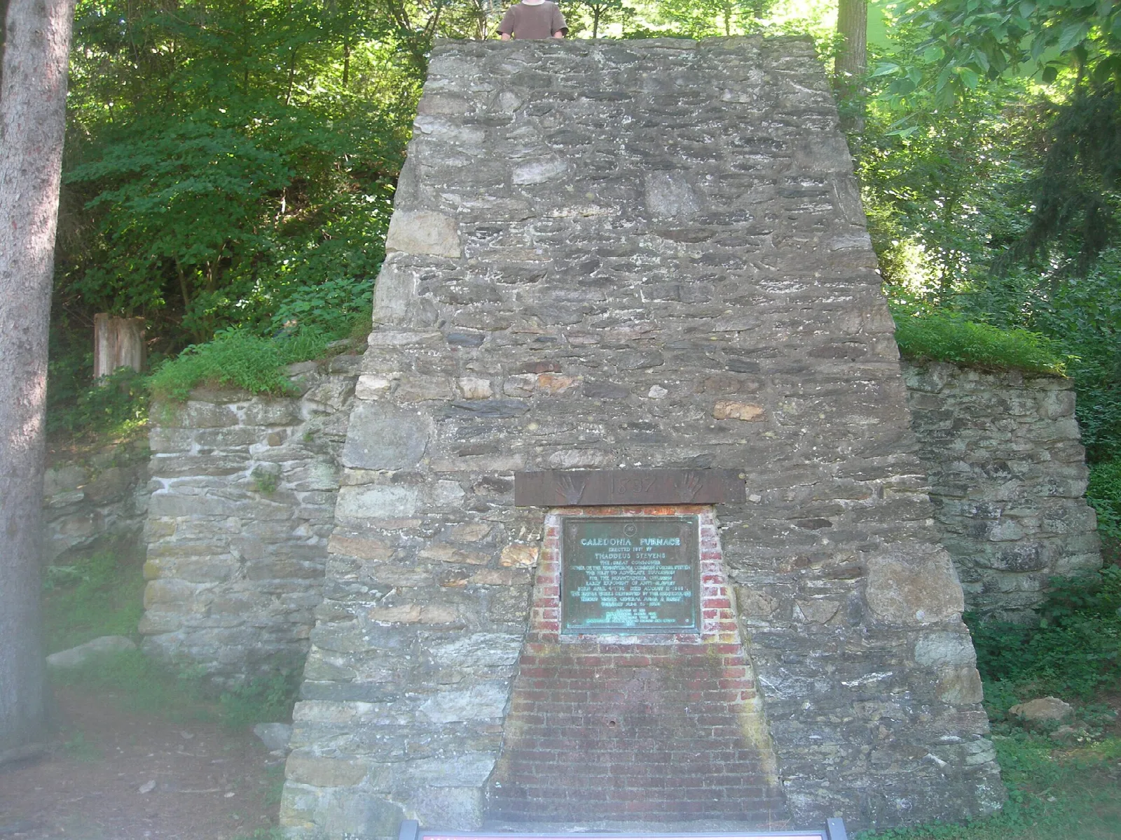 Tall stone furnace stack with a plaque, surrounded by trees
