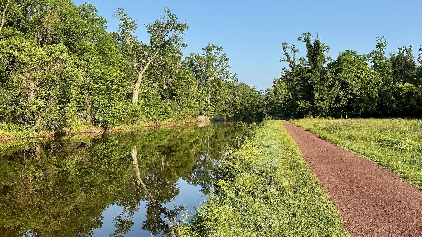 Still canal reflecting trees beside a reddish towpath and grass