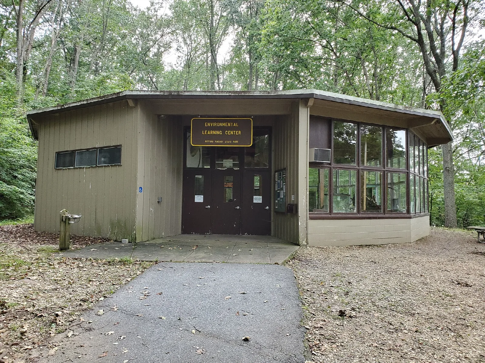 Low tan building with large windows and a paved path in front, among trees