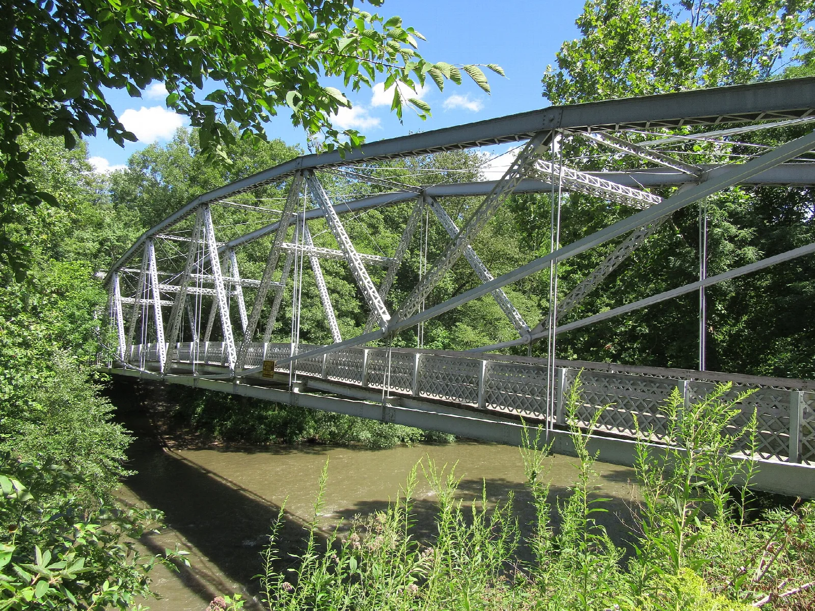 Steel arch truss bridge over a muddy creek with green plants on the bank