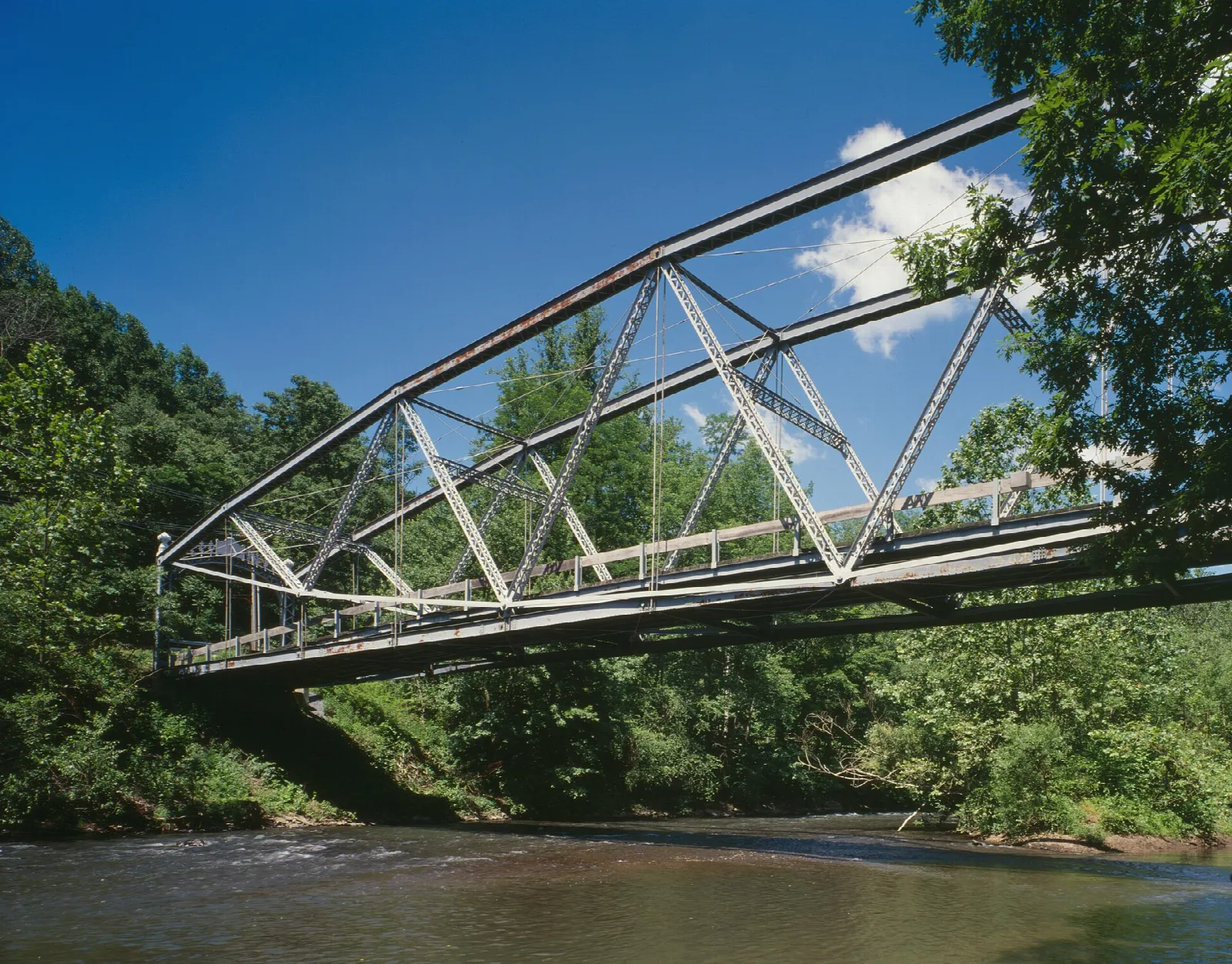 Steel truss bridge spanning a wide brown creek with wooded banks