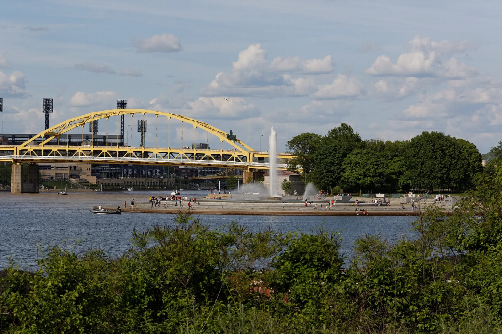 Fountain spraying beside a river with a yellow arch bridge behind