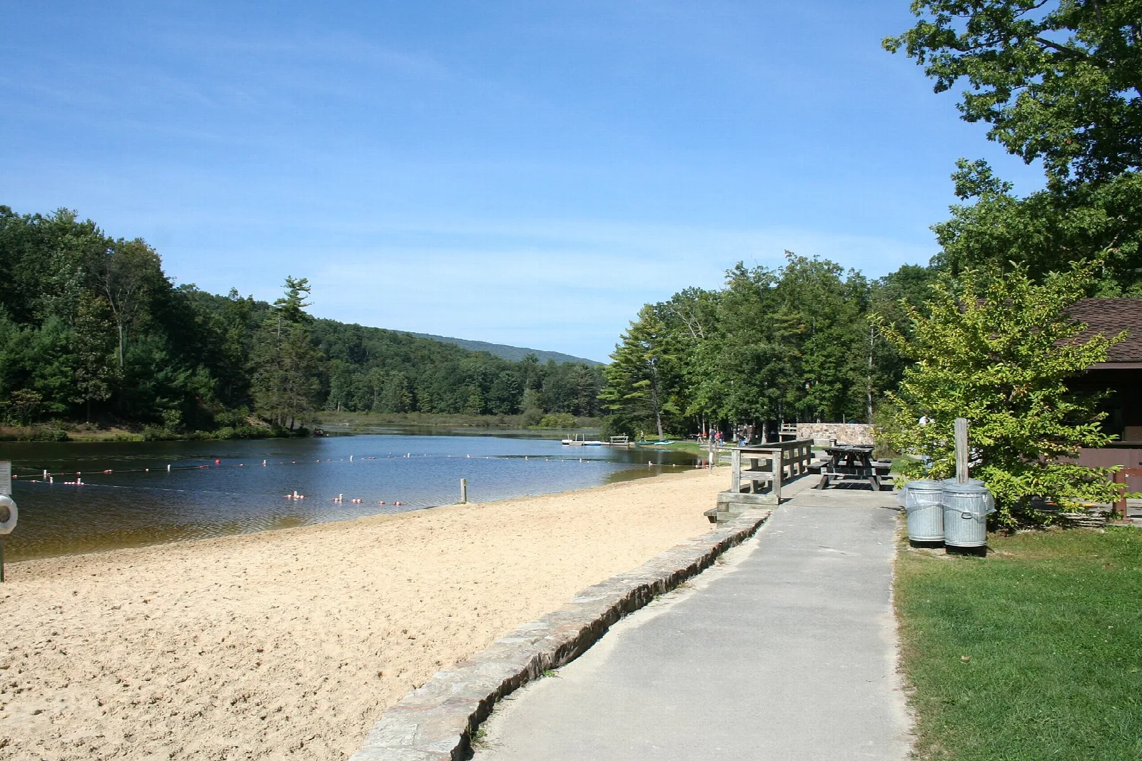 Sandy beach and paved path beside a lake with wooded hills beyond