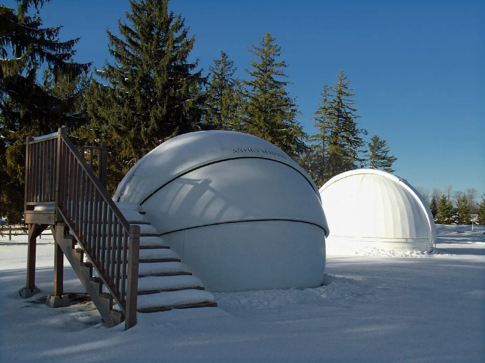Two white observatory domes beside a snowy staircase and evergreens