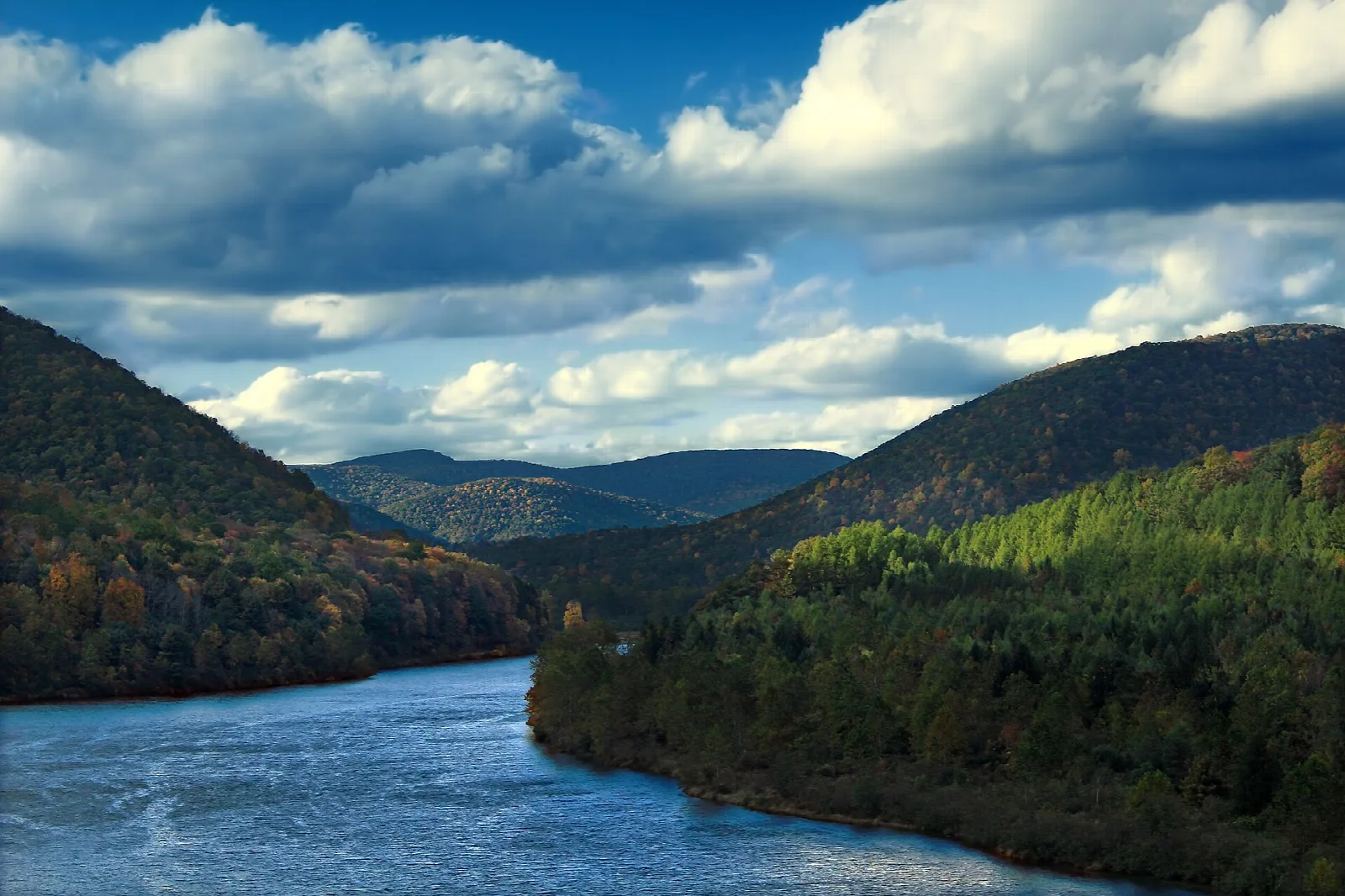 Blue reservoir winding between forested mountains under puffy clouds