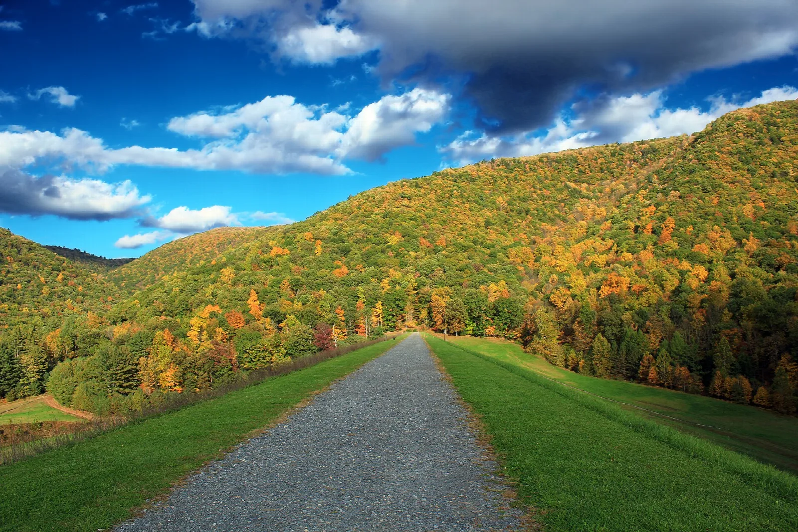 Gravel path across a grassy dam crest toward a fall-colored hillside