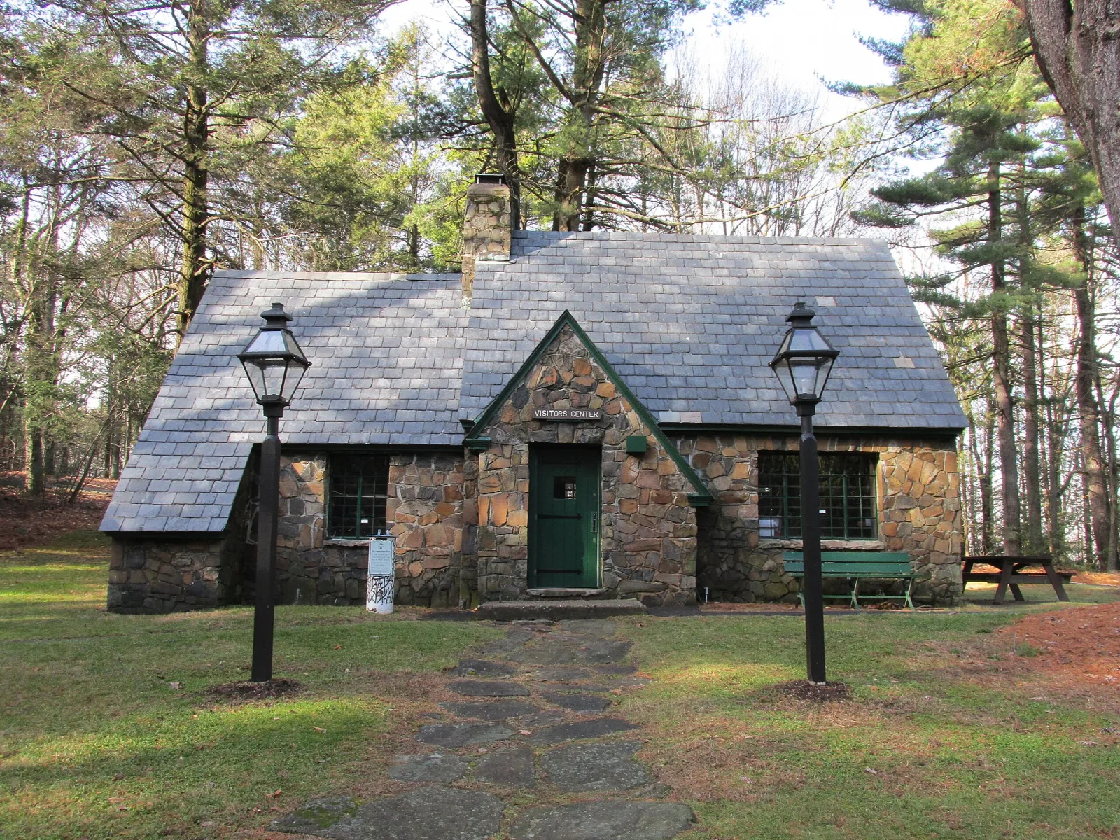 A small stone cottage with a slate roof and two lamp posts on a lawn among pines