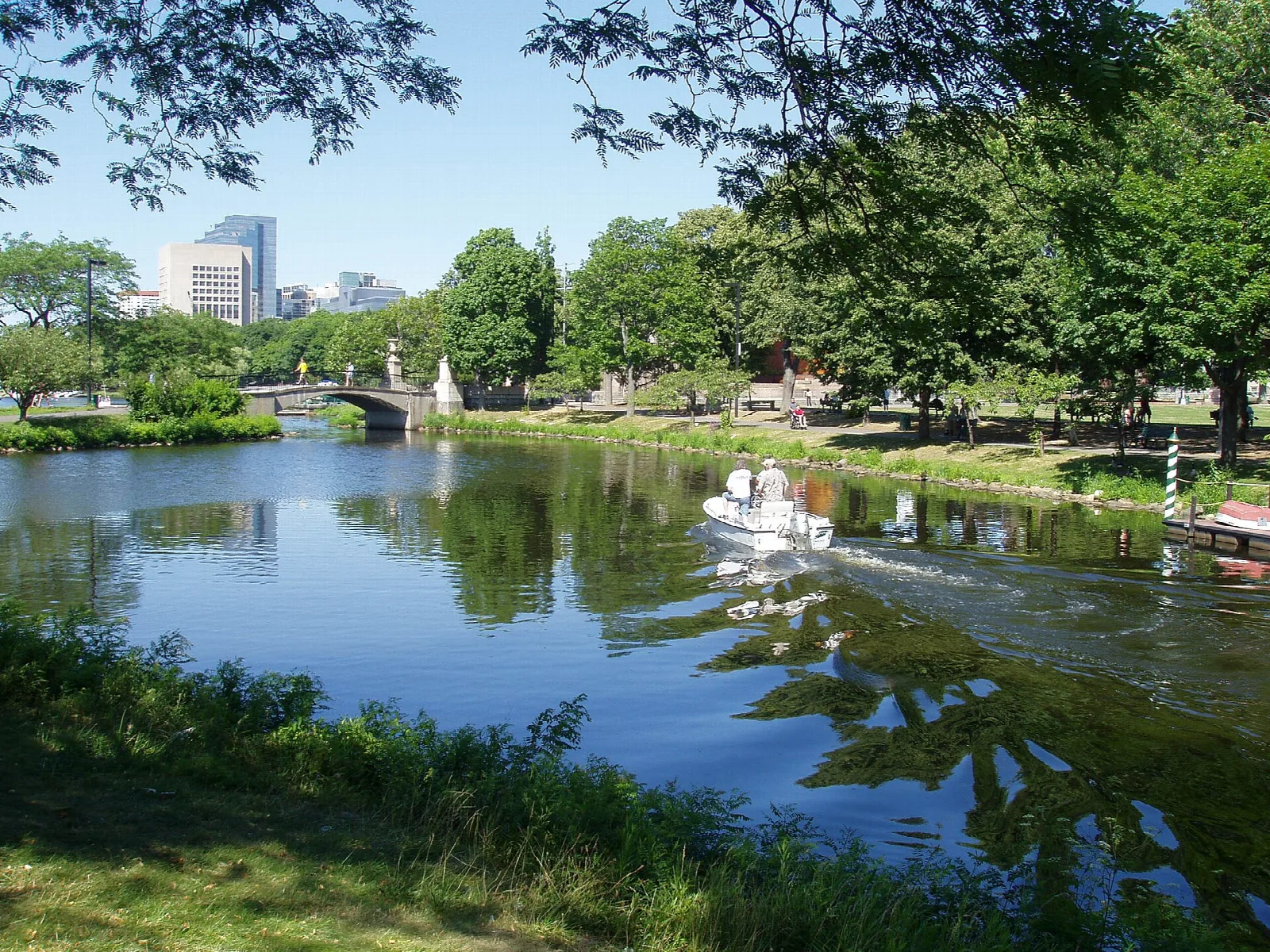 A boat on the Charles River beside a grassy bank, with a bridge and city buildings far off