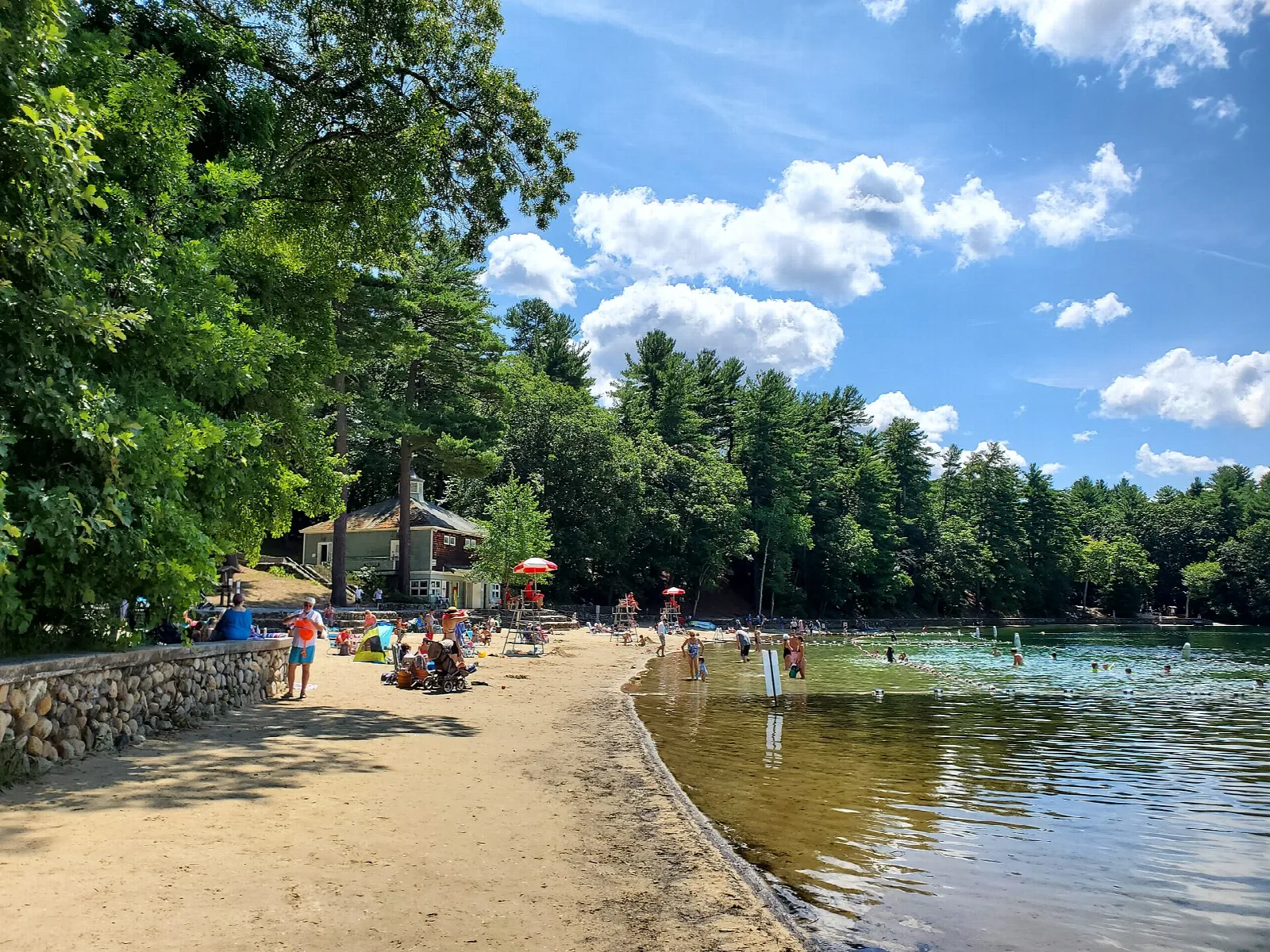 A sandy beach with swimmers and a small shelter beside the lake under blue sky