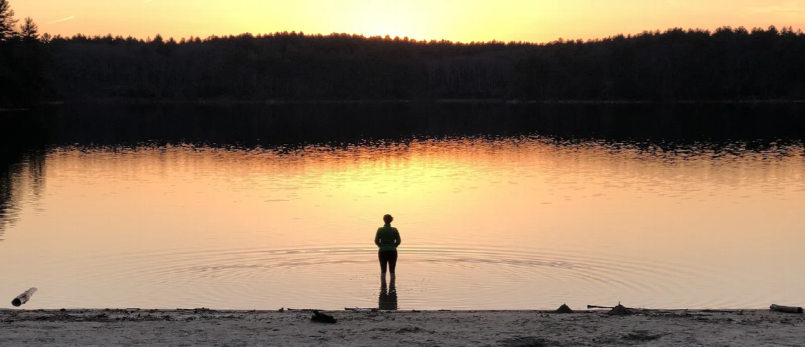 A person standing at the water's edge at sunset on a still lake
