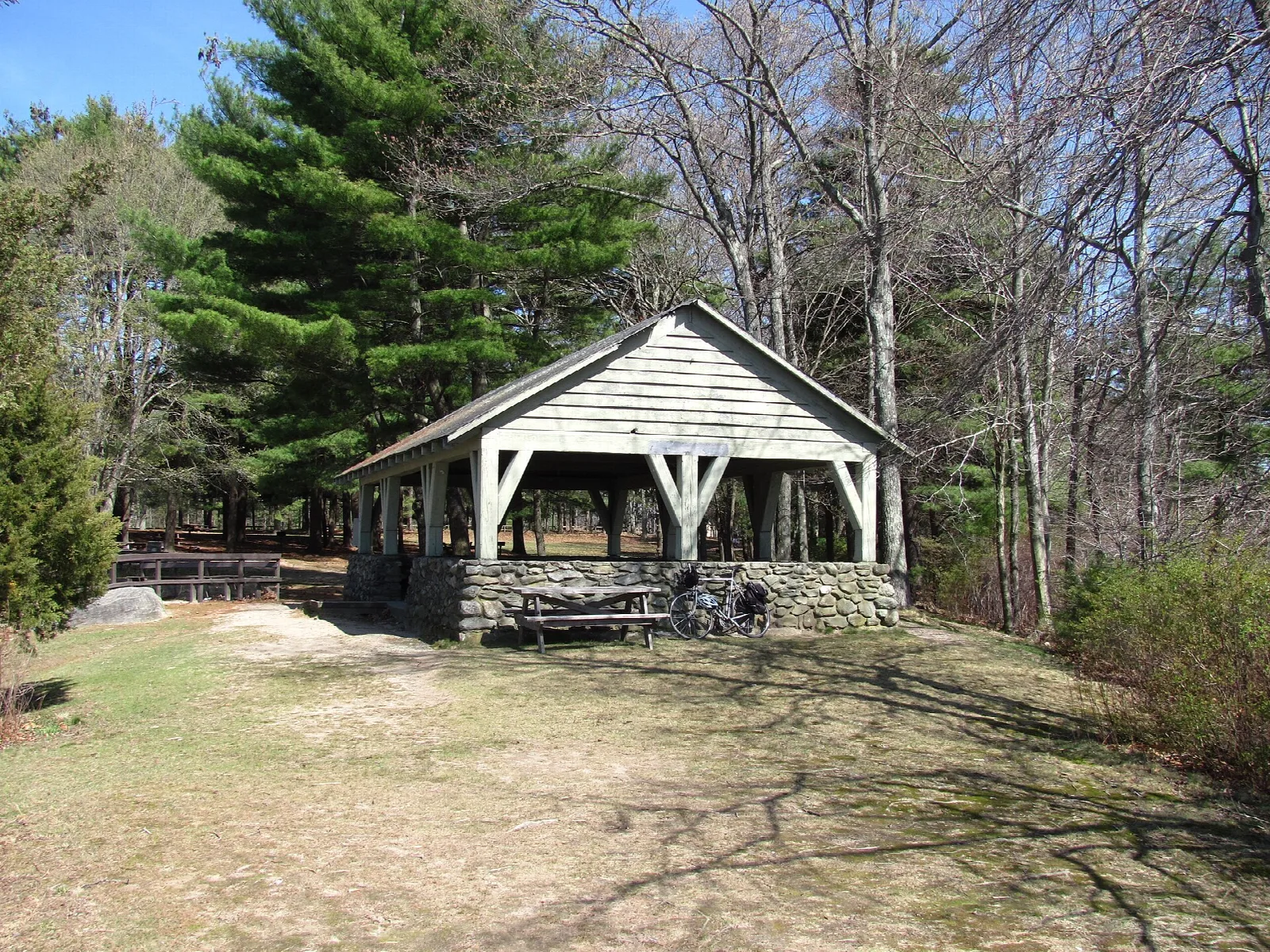 An open wooden pavilion with a stone base among pines and bare trees