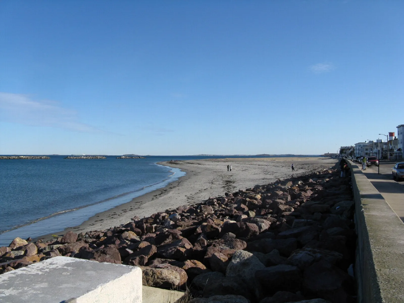 A sandy beach beside a seawall of large rocks, under blue sky