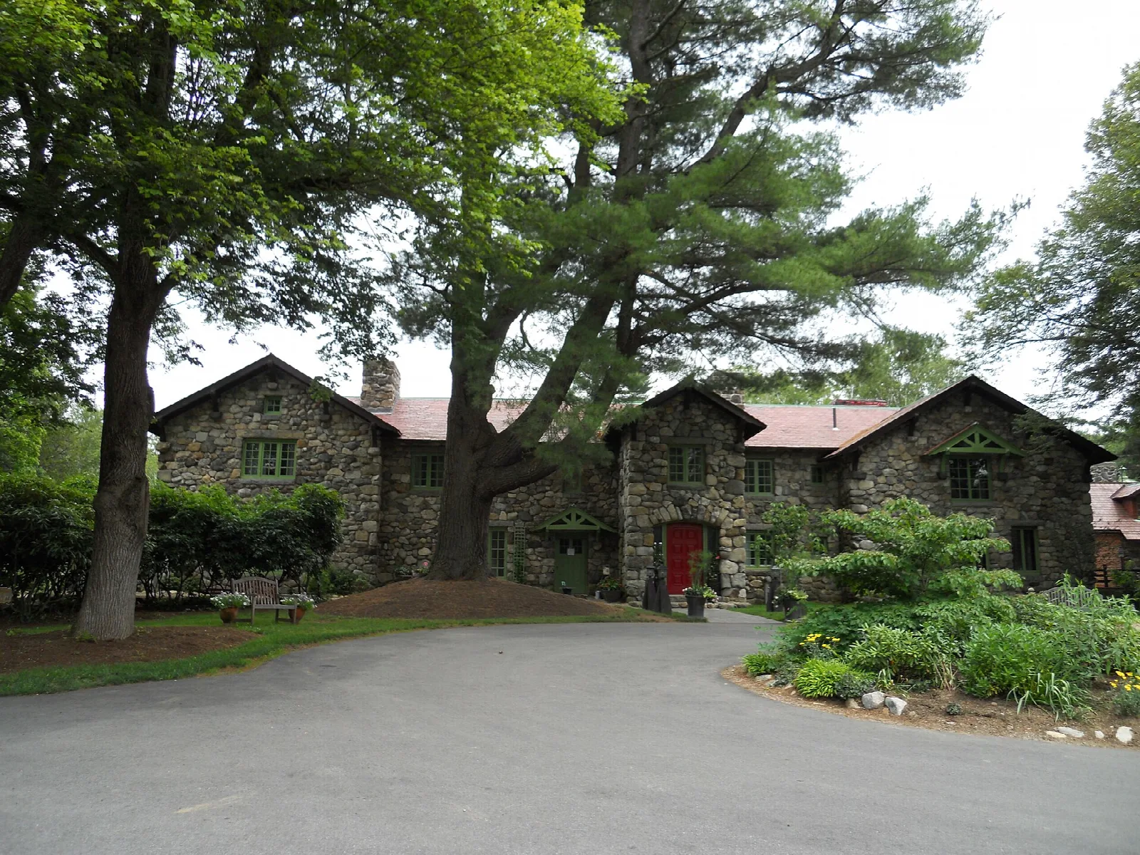 A stone house with a red door and a paved drive among tall trees