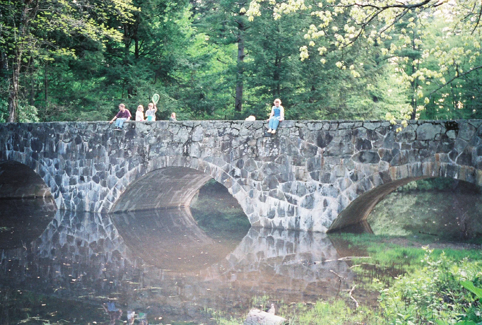 A stone arch bridge over still water with trees behind and people on top