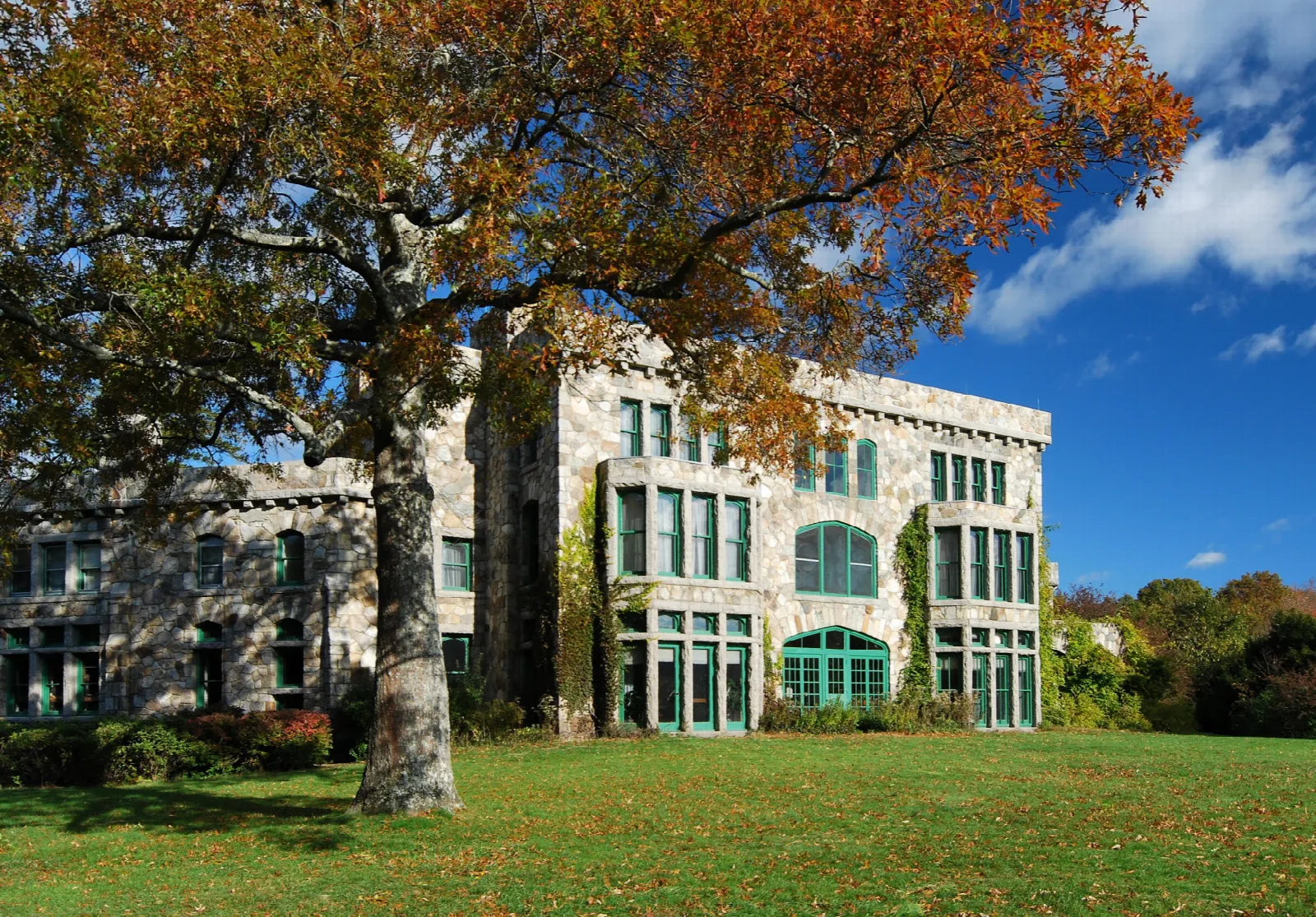 A large stone mansion with big windows beside an autumn tree on a lawn