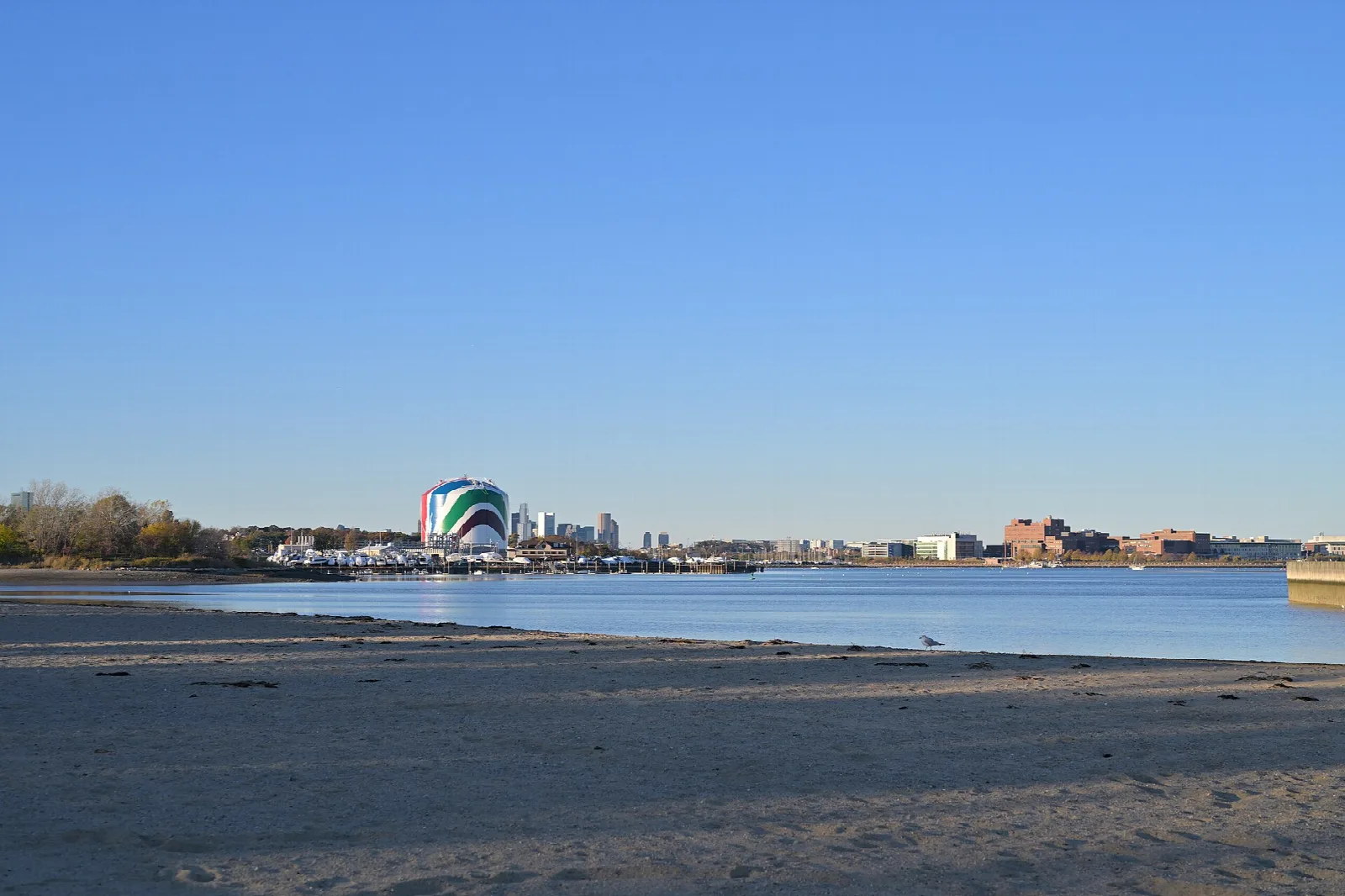 A sandy shore looking across calm water toward a distant striped tank and city skyline