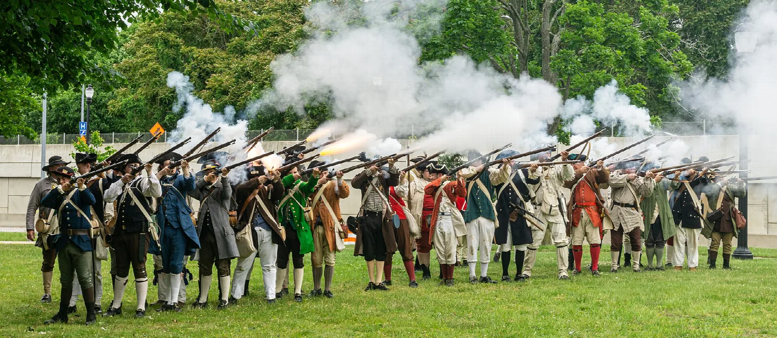 Costumed reenactors firing muskets in a grass field with white smoke and trees behind