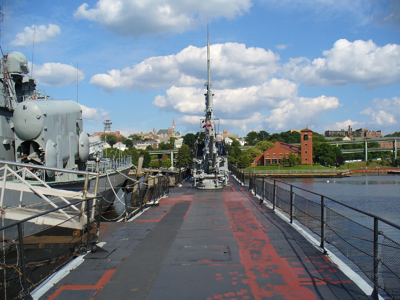 Gray battleship deck with red markings and gun mounts, a brick tower on the far shore