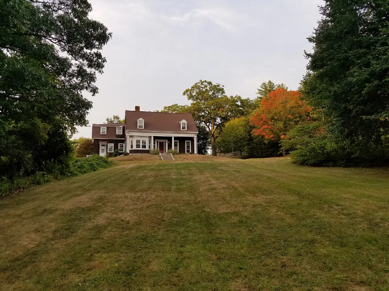 Two-story house with a dark roof across a lawn, with fall trees behind