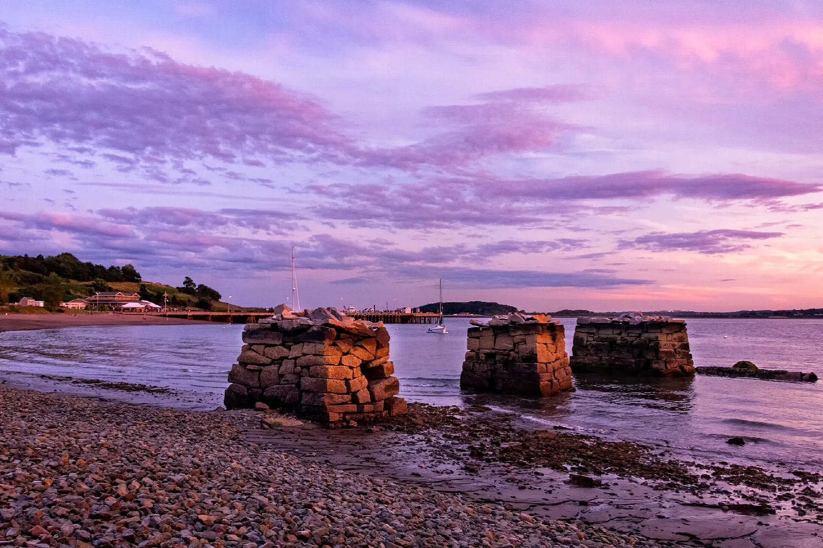 Old stone pier remains on a pebble shore under a pink and purple sunset sky