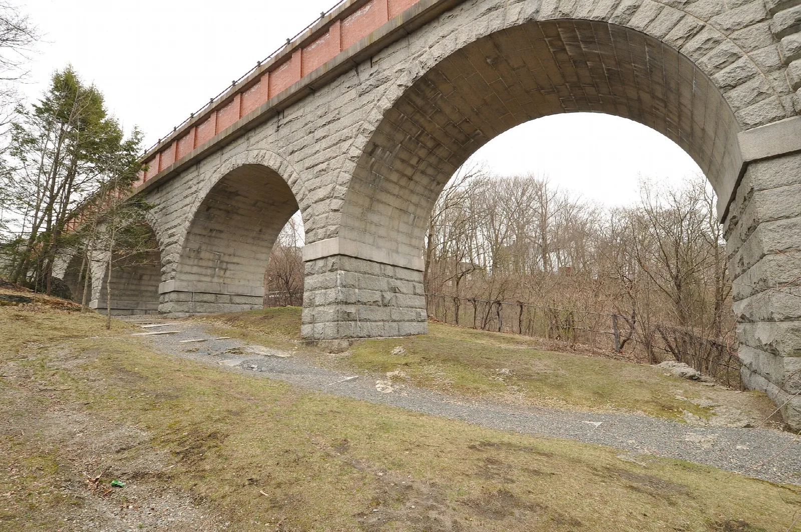 Stone arches of a bridge with a red rail on top, seen from a gravel path