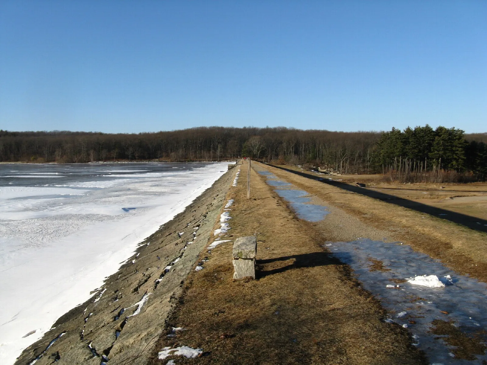 Stone-faced dam crest with a path and patches of snow, woods beyond