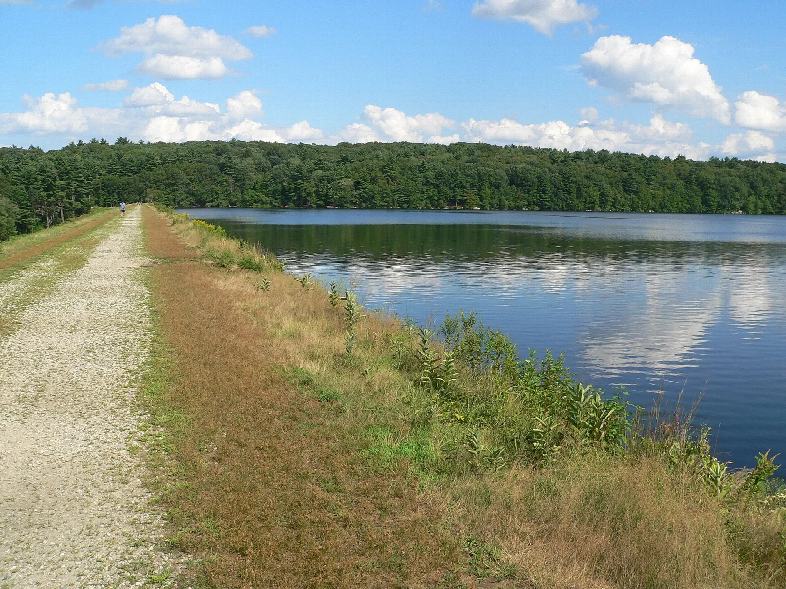 Gravel dam path beside a calm reservoir and forest under blue sky