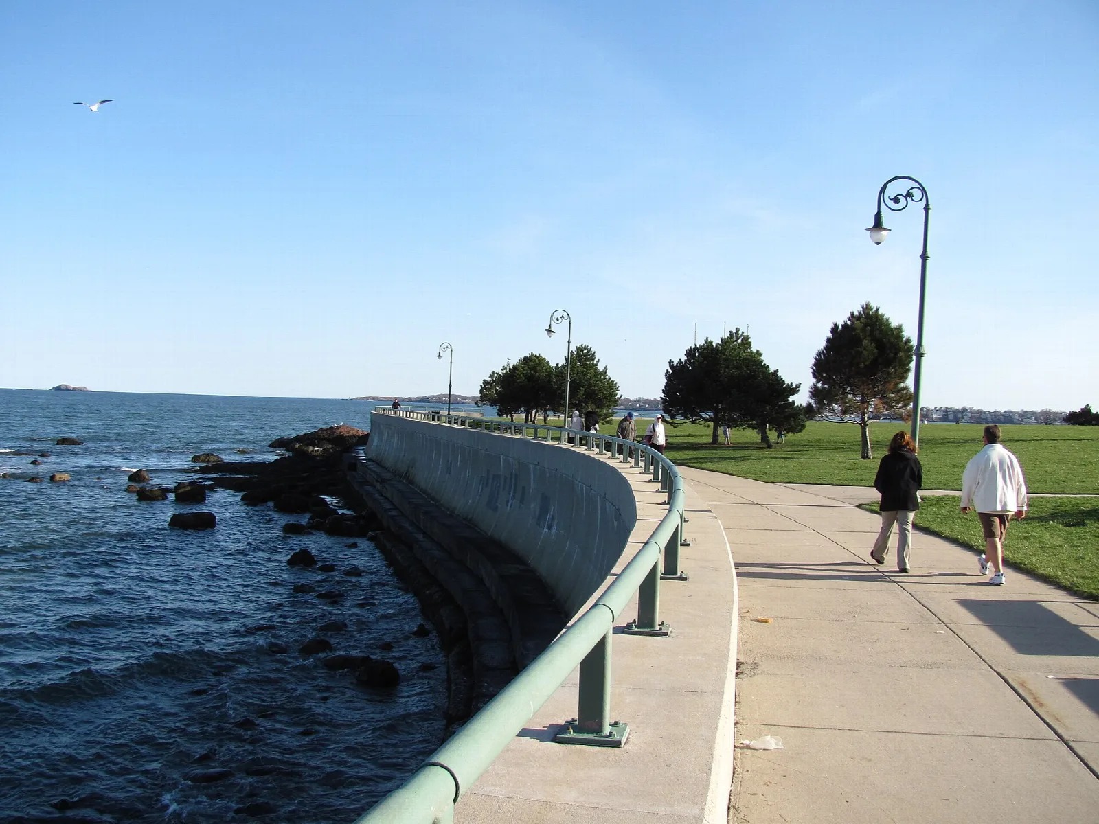 Seawall walkway along rocky shore with two people walking beside a grassy park