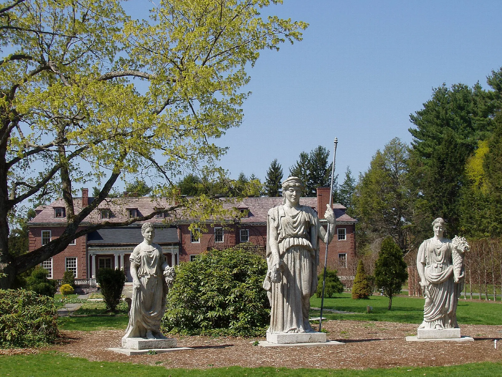 Three white statues in a garden in front of a brick mansion