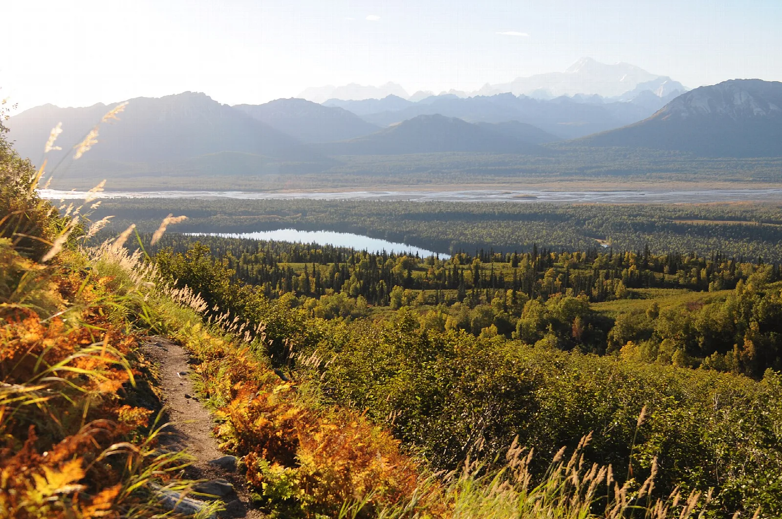 Trail along a golden autumn hillside above a forested valley and lake, with hazy mountains beyond