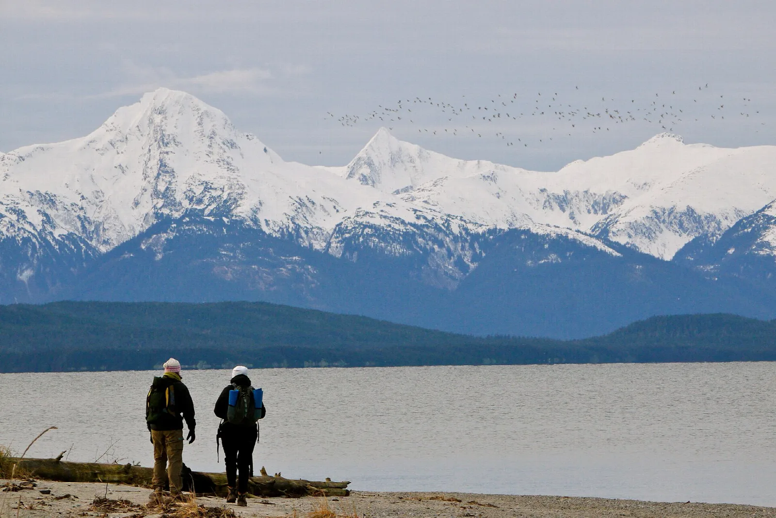 Two people on a gravel shore looking at a snow-capped mountain range across the water