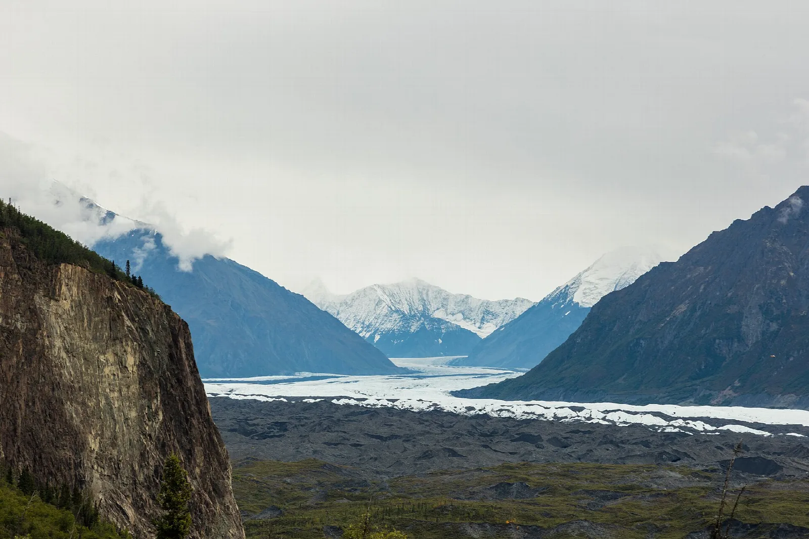 Glacier edge below a steep rock wall, with clouded mountains in the distance
