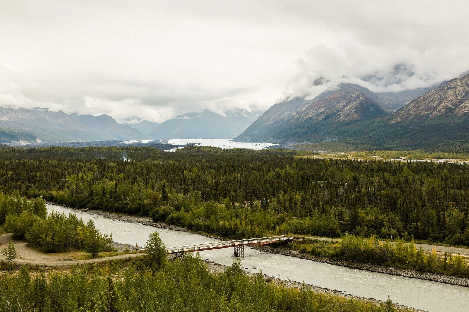 Braided river and a bridge in a forested valley, with a glacier and cloudy mountains beyond