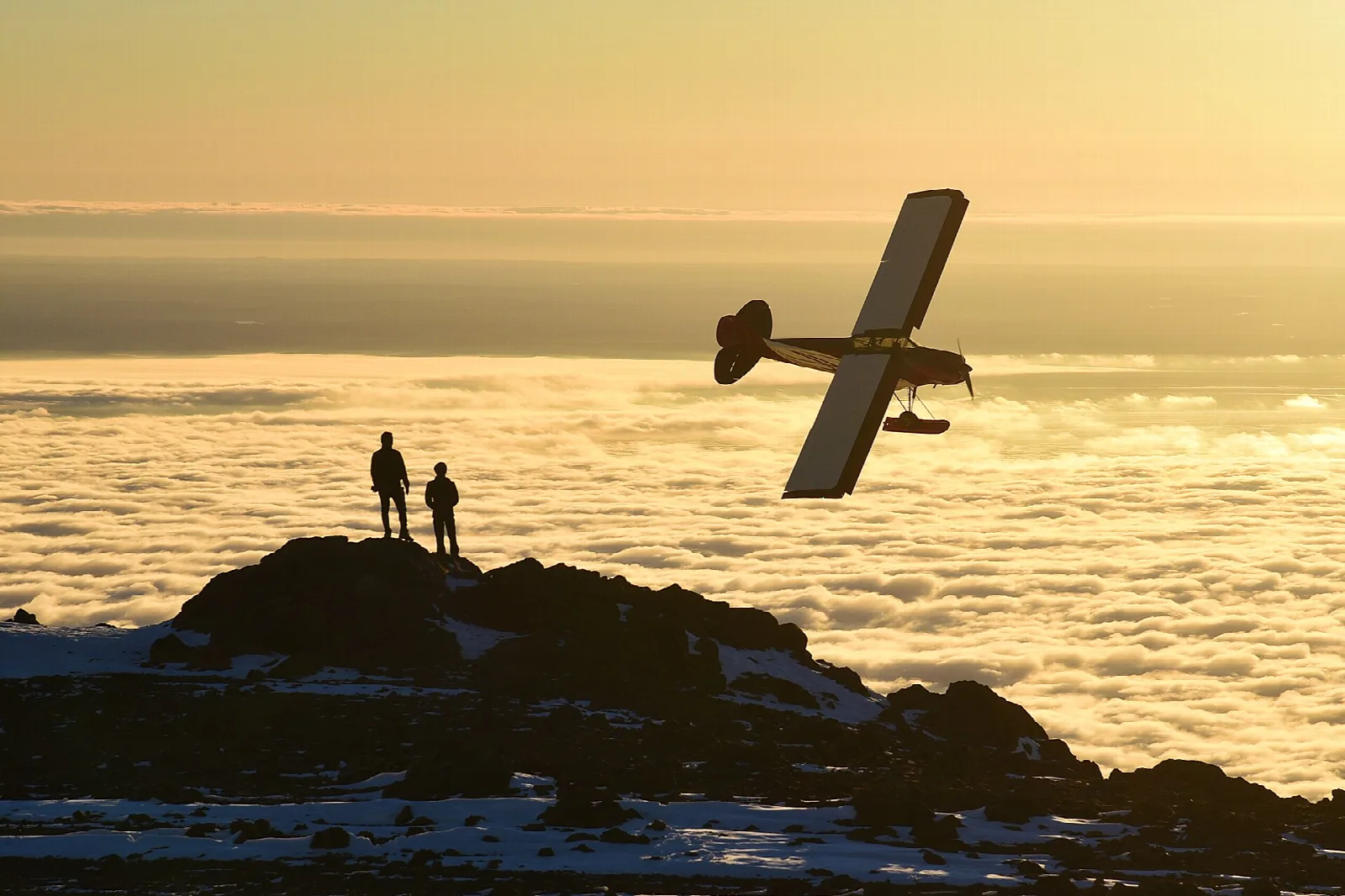 Two people on a rocky summit watching a small plane above a sea of cloud