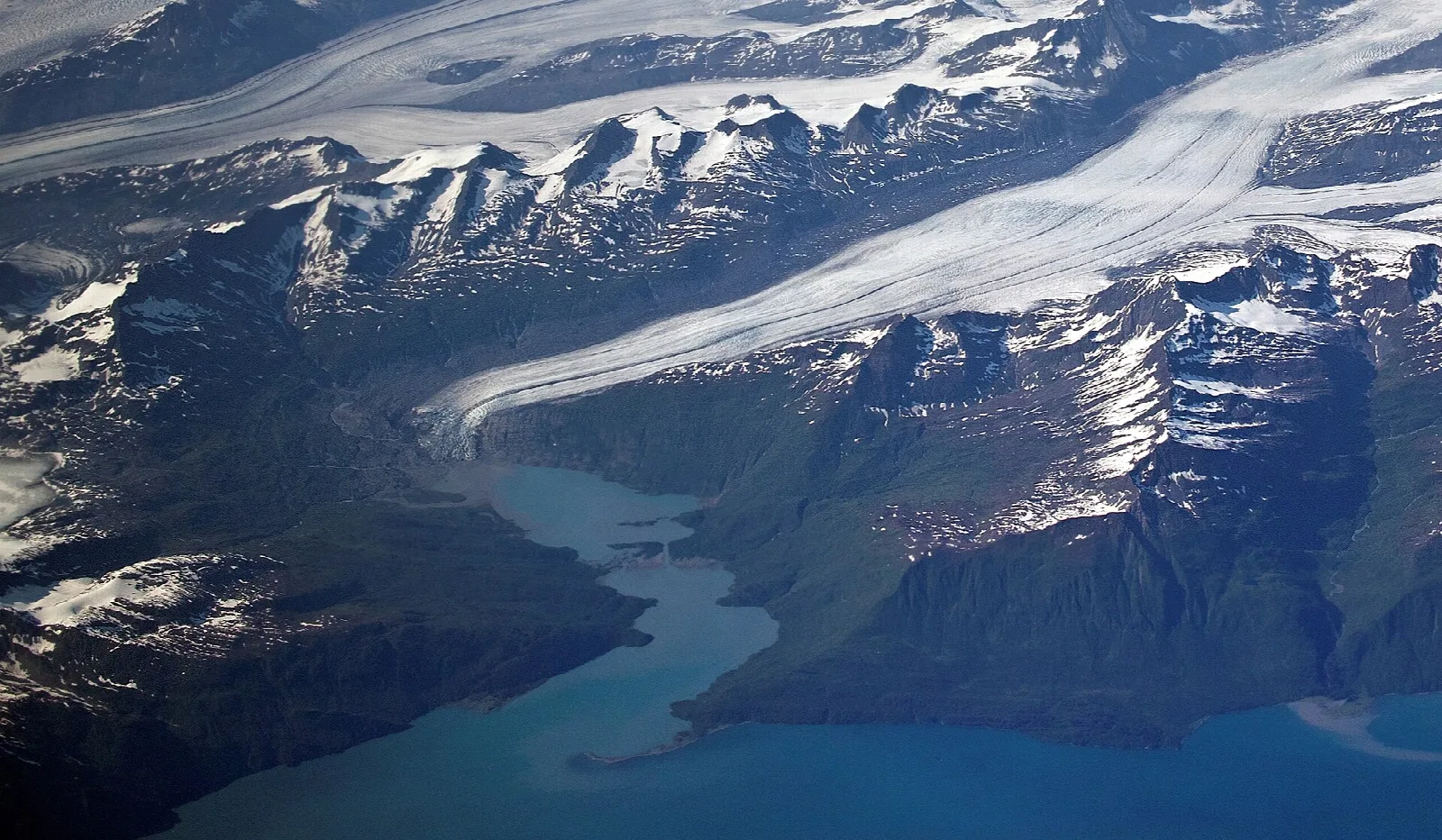 Aerial view of a glacier flowing between snowy mountains toward blue-green water