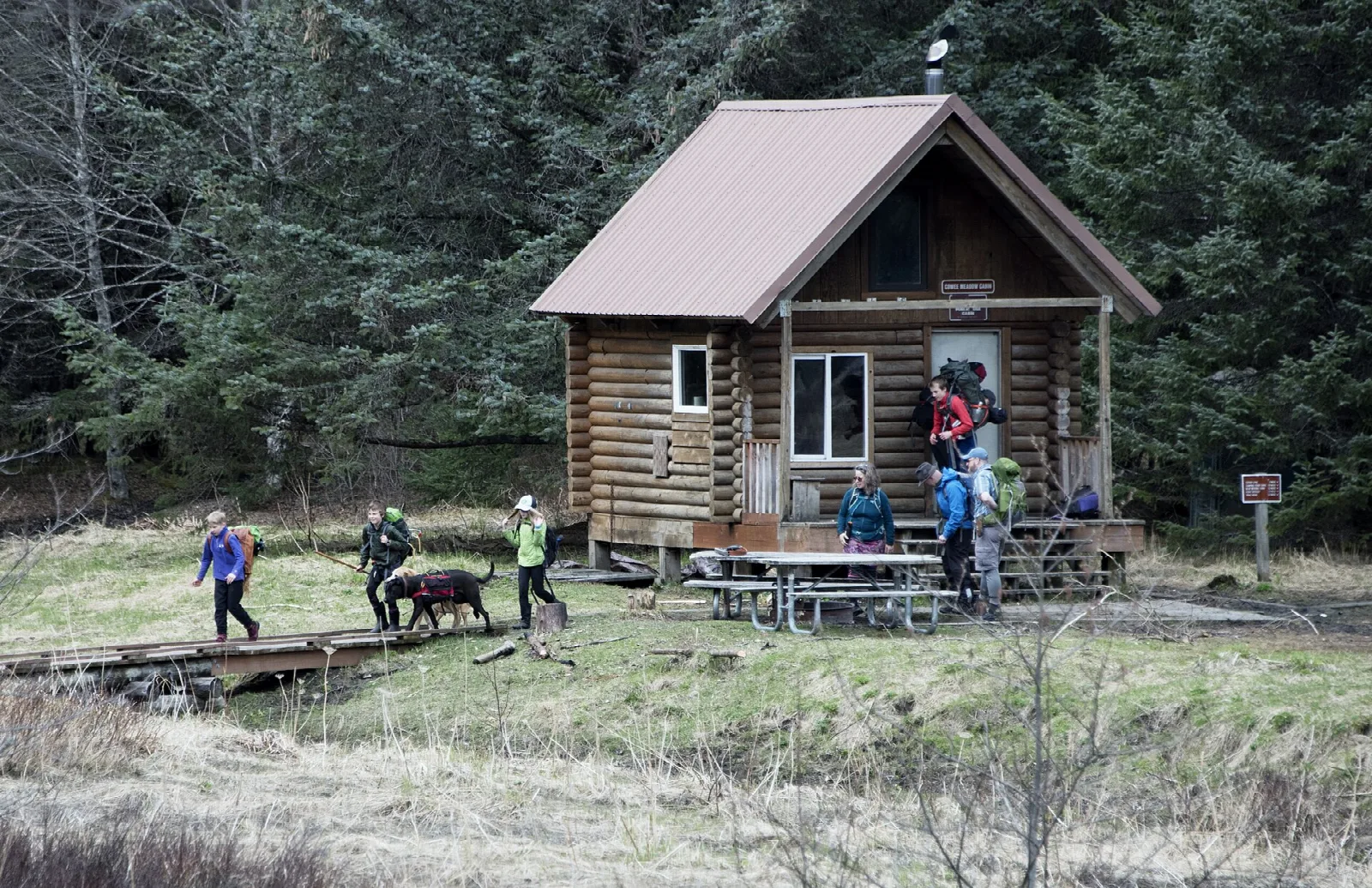 Hikers and a dog gathered on the porch of a small log cabin in a meadow