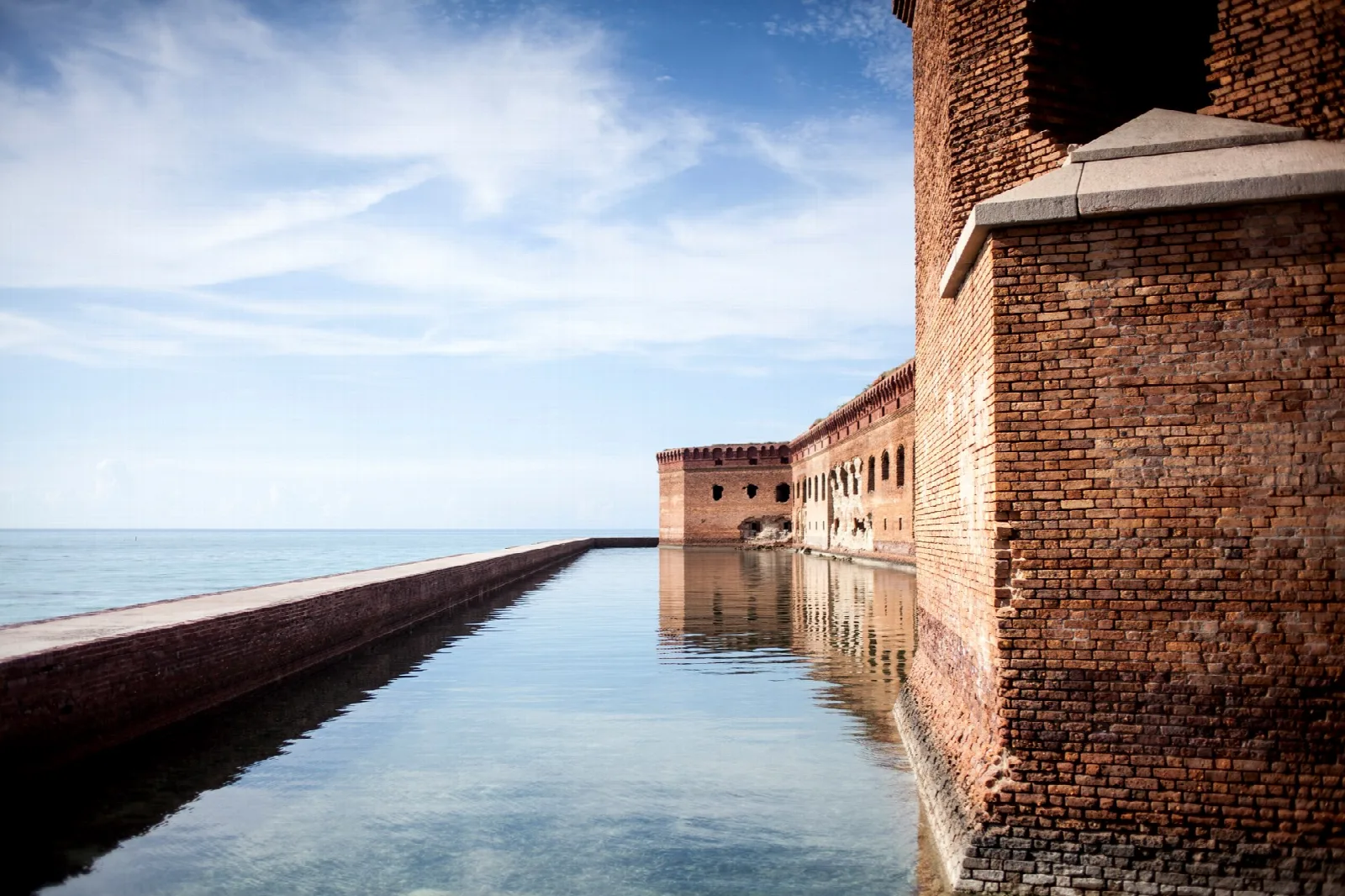 A narrow seawall running along the fort's brick wall through calm, clear water