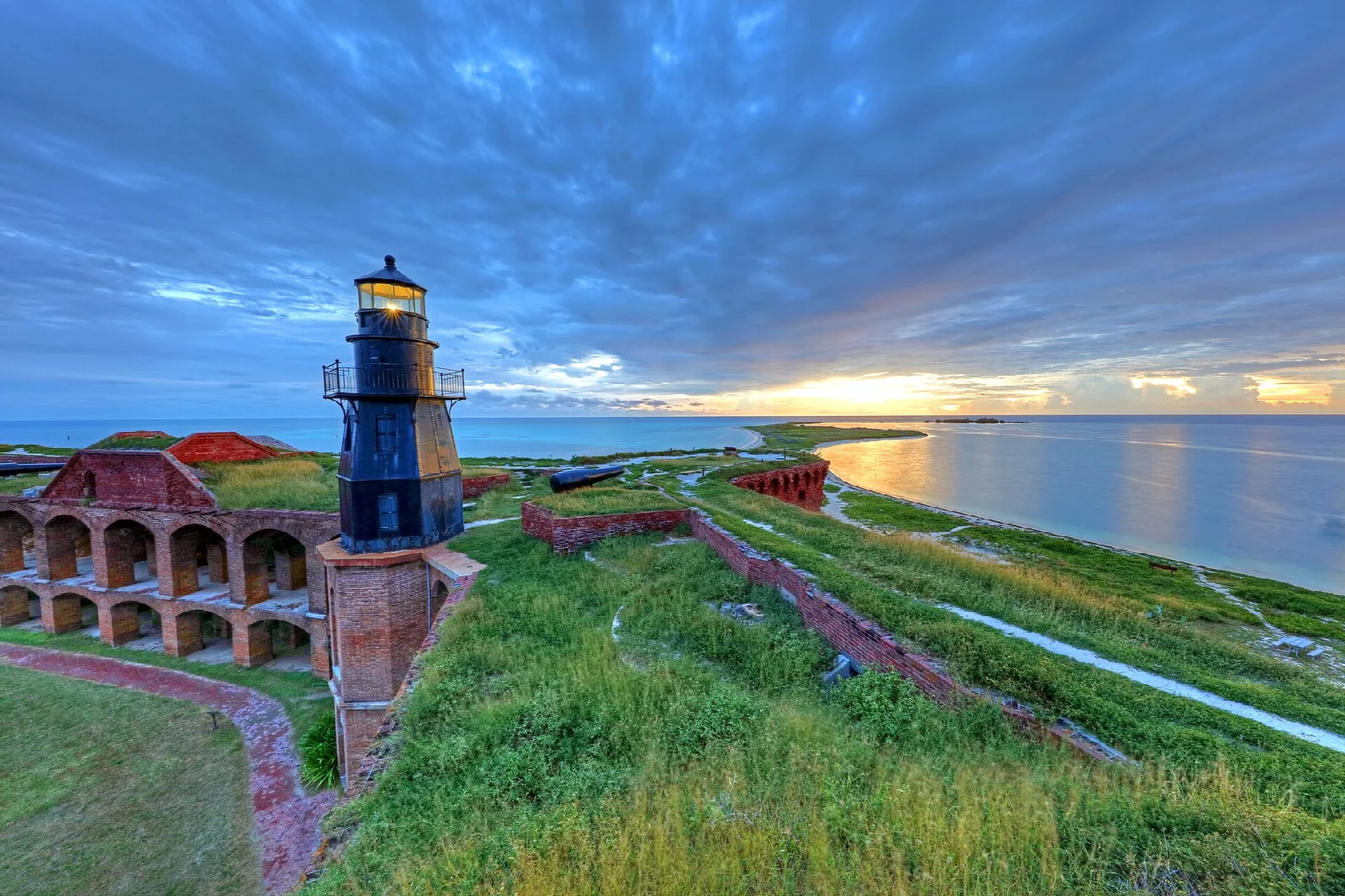 A black lighthouse atop the fort's grassy rampart at sunset over the sea