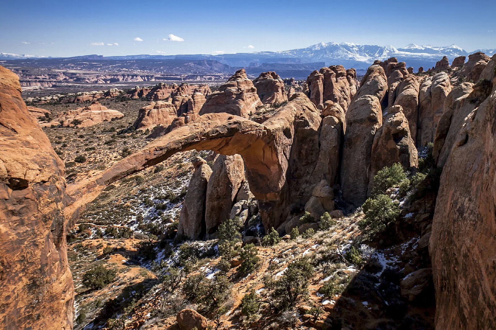 A long, thin stone arch spanning a sandstone valley with snowy mountains in the distance