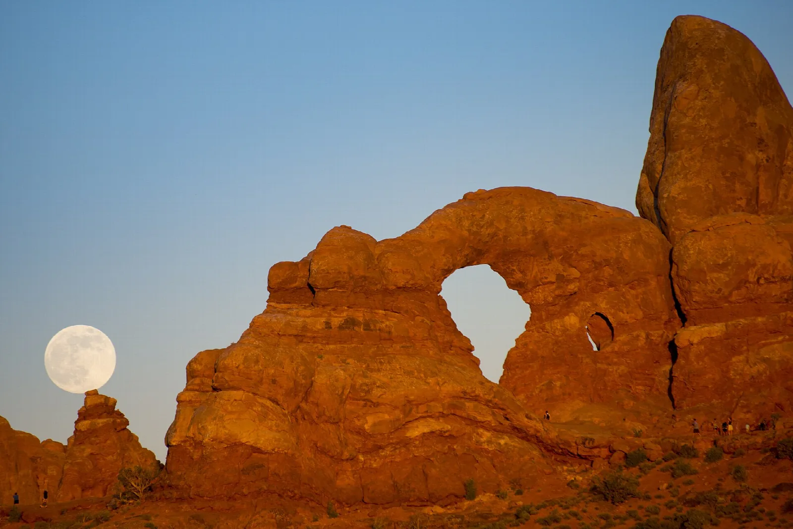 Full moon low in a blue evening sky beside a sunlit sandstone arch and tower