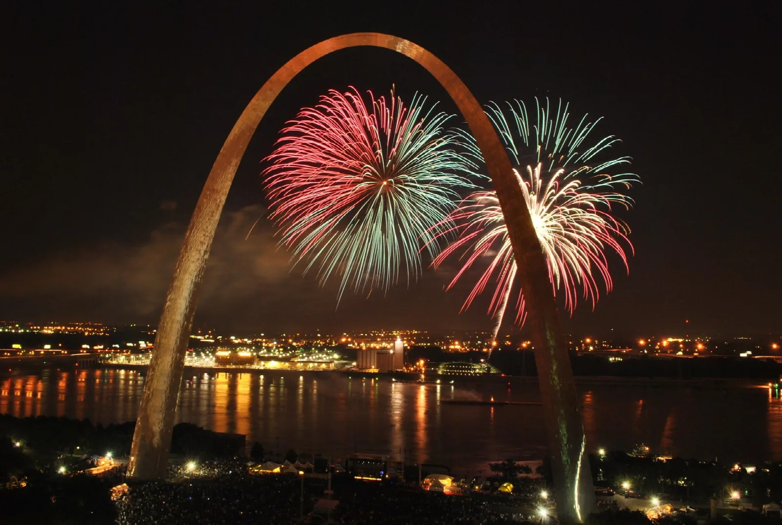 Red and green fireworks bursting through the lit arch above the riverfront at night