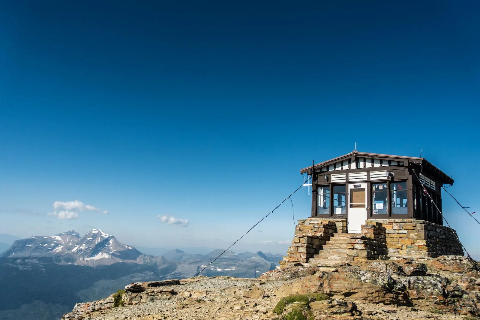 A small, brown and white structure sits on a rocky top with mountain peaks in the distance.