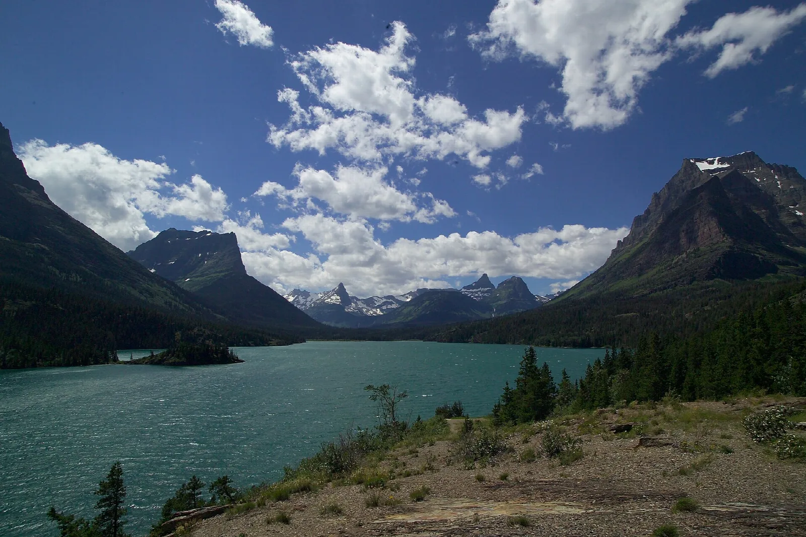 A small forested island in a long blue lake between steep mountains under puffy clouds