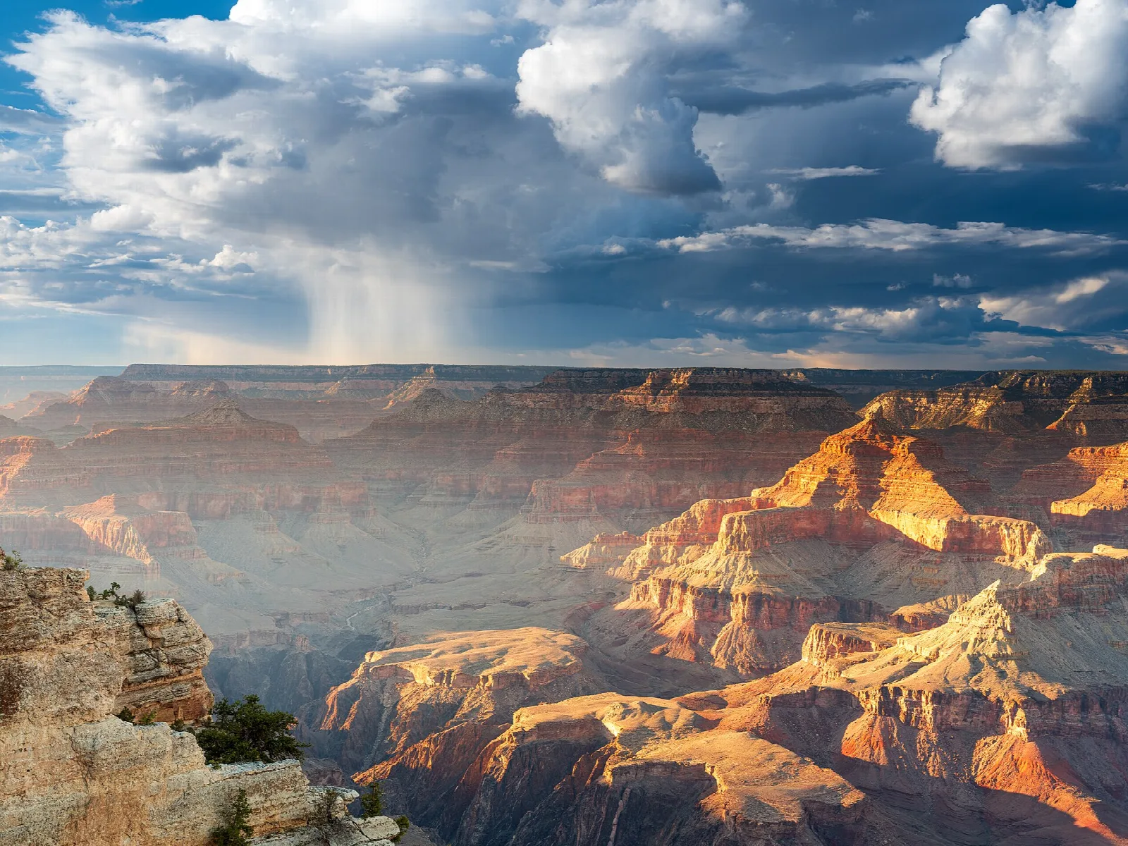 A rain shaft falling from storm clouds over sunlit buttes and canyons