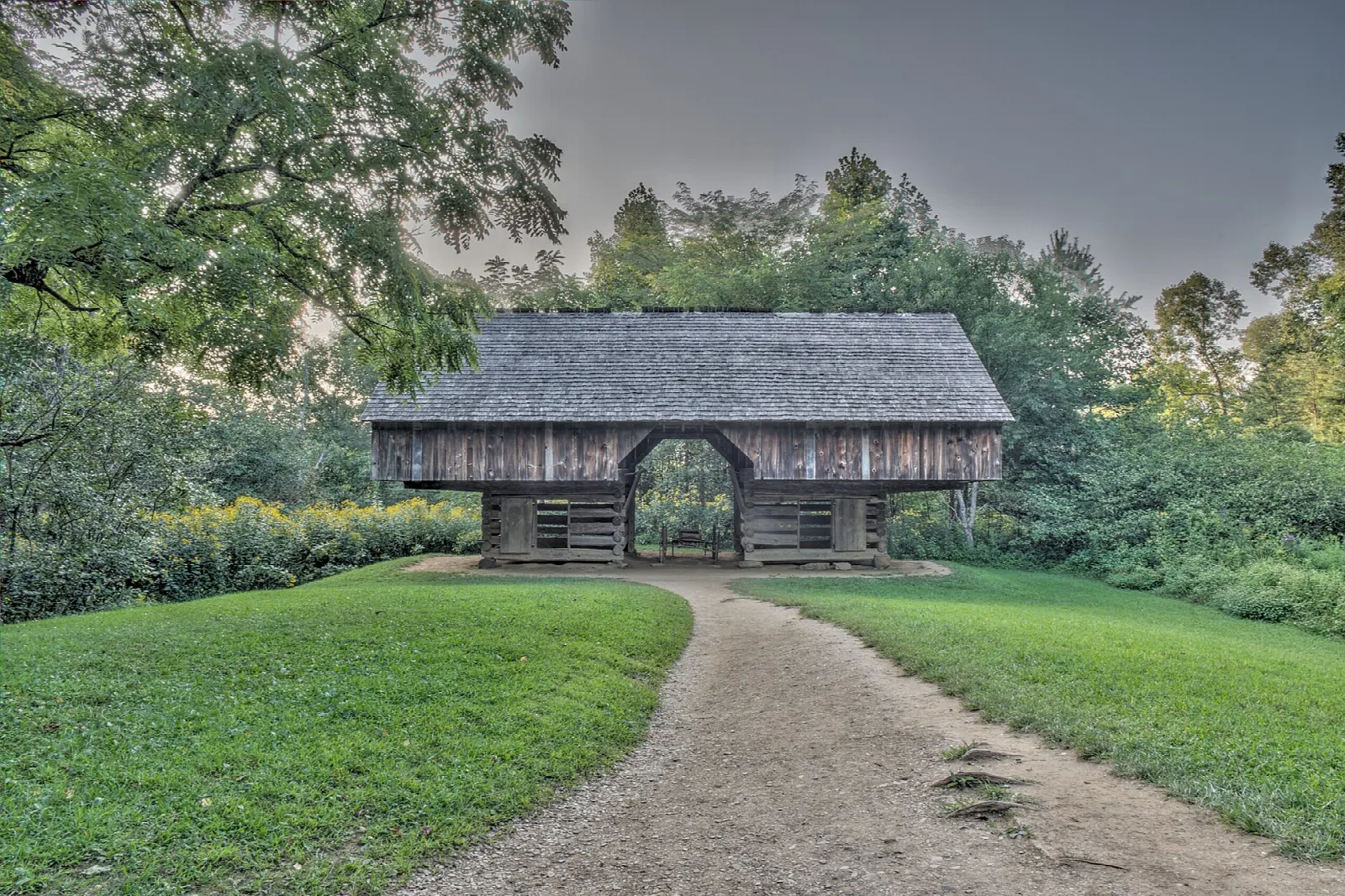 A historic log barn with an overhanging loft at the end of a gravel path through green lawn