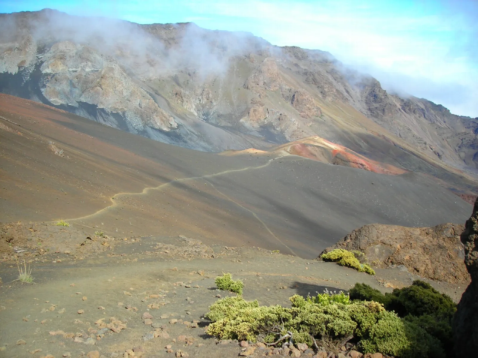 A trail switchbacking down into a vast, colorful volcanic crater with clouds on the rim