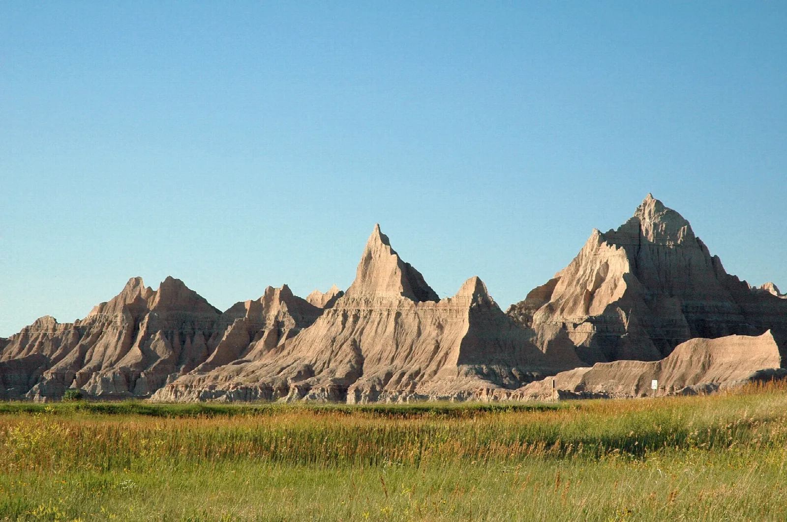 Row of sharp, sunlit badlands spires rising above green grassland under a clear sky