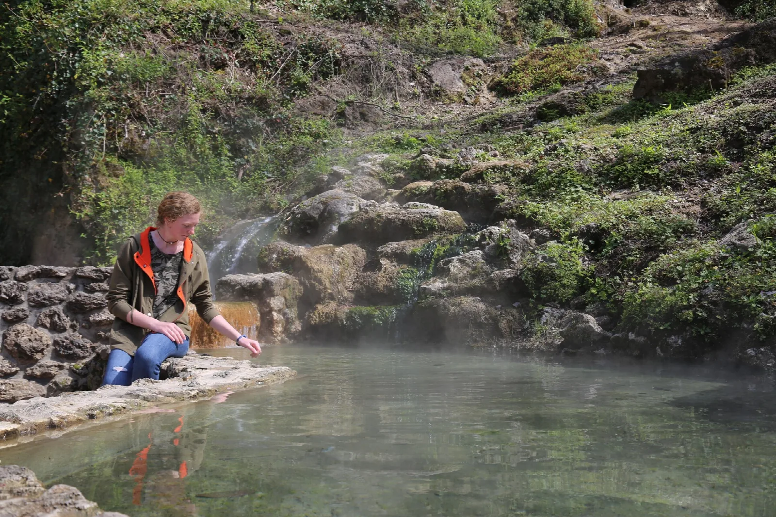 A visitor touching steaming thermal water flowing over mossy rock into a pool